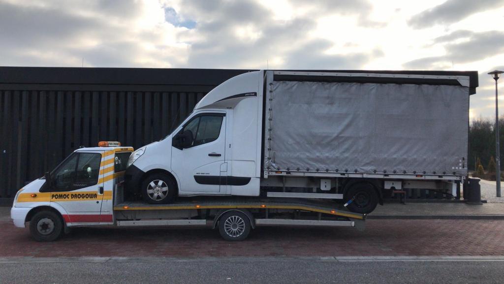 Laweta z pomarańczowym kogutem przewozi białego busa z plandeką na tle ciemnego budynku.