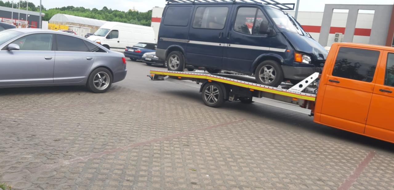 Uszkodzony, granatowy bus z bagażnikiem dachowym na lawecie, pomarańczowy holownik, w tle szary samochód osobowy i biały dostawczak na parkingu.