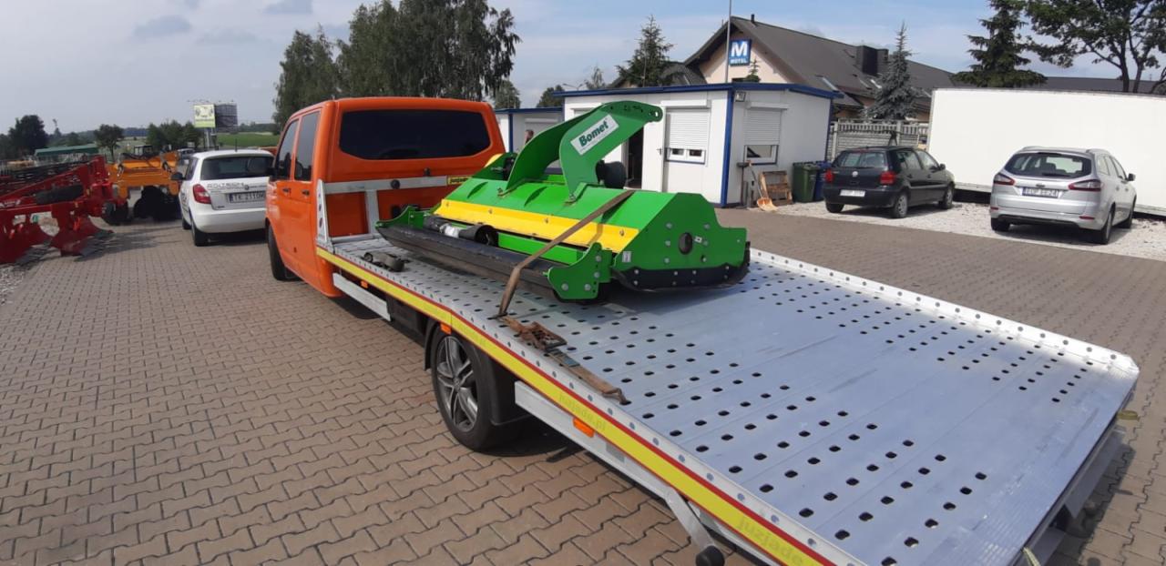 Pomarańczowy bus z platformą transportową przewozi zielono-żółtą kosiarkę marki Bomet na tle placu z kostki brukowej i budynków.