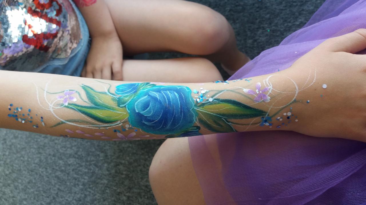 Ręka dziecka z namalowaną niebieską różą i brokatem, otoczona drobnymi kwiatkami i białymi ornamentami.