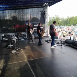 Zespół muzyczny gra koncert plenerowy w Katowicach, widoczni gitarzyści, saksofonista oraz perkusja na scenie, w tle publiczność.