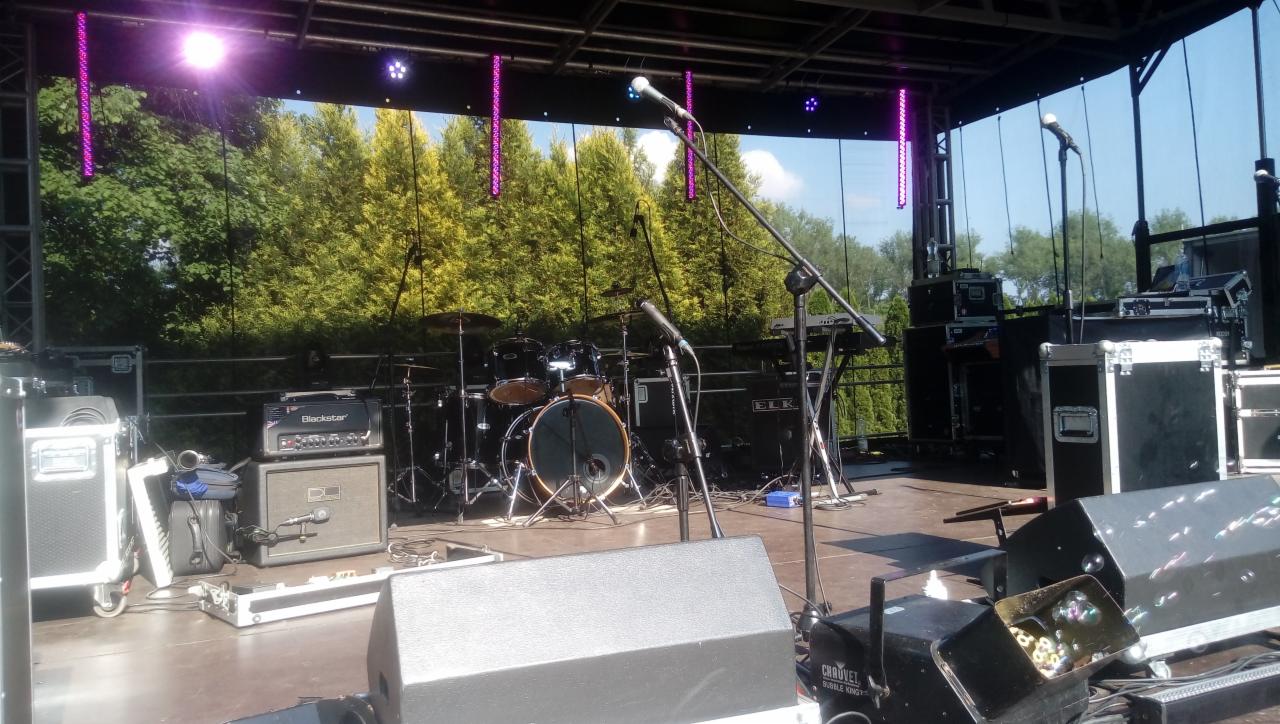 Scena plenerowa z zestawem perkusyjnym, wzmacniaczami Blackstar i ELK, klawiszami oraz mikrofonami, oświetlona fioletowymi lampami, z zielonymi drzewami w tle.