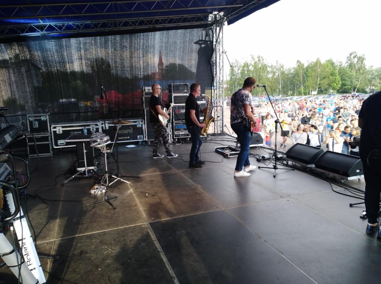 Zespół muzyczny gra koncert plenerowy w Katowicach, widoczni gitarzyści, saksofonista oraz perkusja na scenie, w tle publiczność.