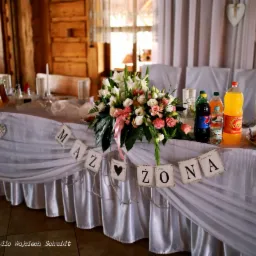 Dekoracja stołu weselnego z białym obrusem, kwiatami w kompozycji z różową wstążką, napojami i transparentami z napisem 'Mąż' i 'Żona' na tle drewnianej ściany i białych zasłon.