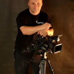 Portret mężczyzny z kamerą Sony na statywie, ubrany w czarną koszulkę z logo 'Digital Studio', na ciemnym tle z bocznym oświetleniem.