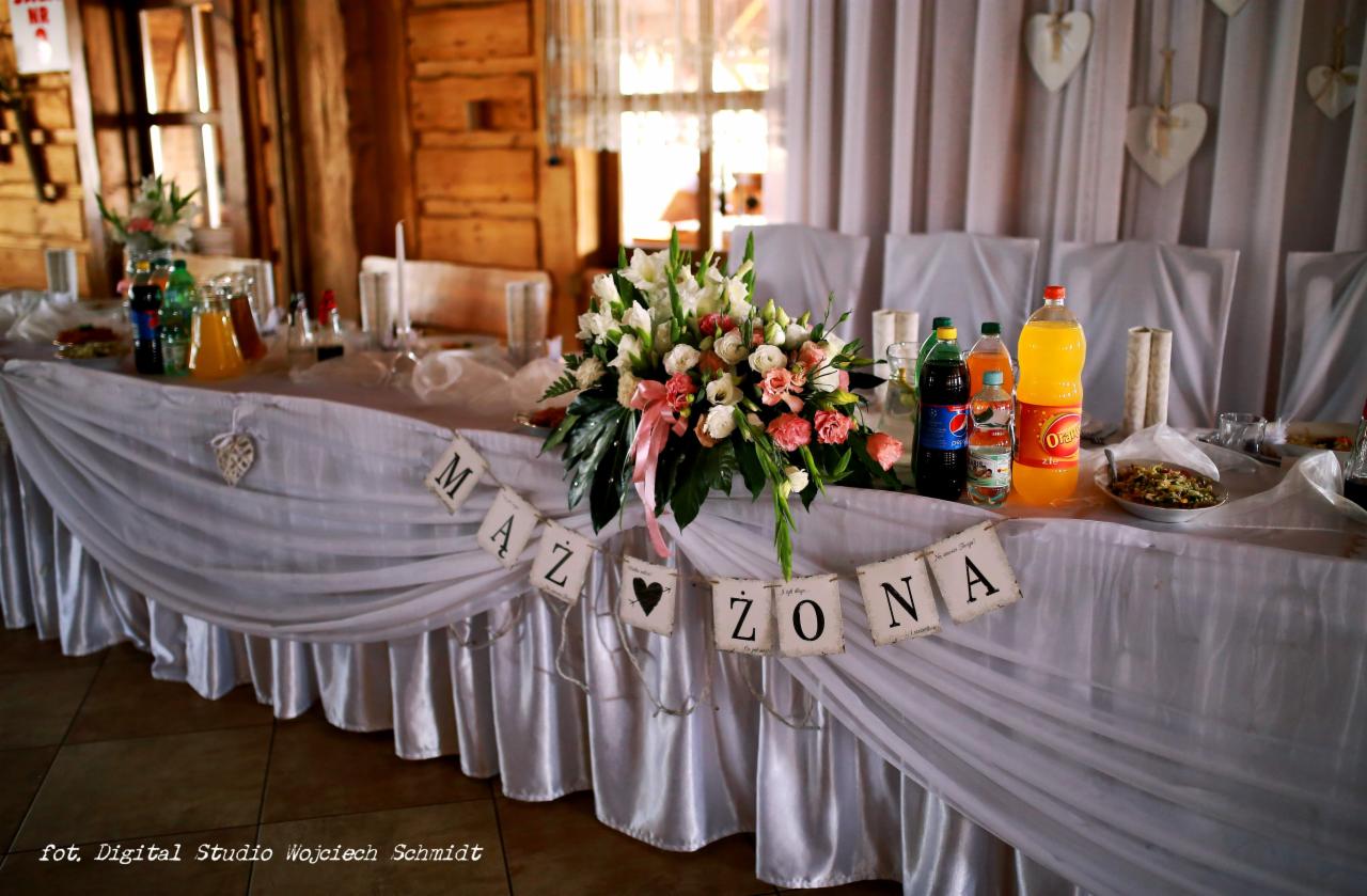 Dekoracja stołu weselnego z białym obrusem, kwiatami w kompozycji z różową wstążką, napojami i transparentami z napisem 'Mąż' i 'Żona' na tle drewnianej ściany i białych zasłon.