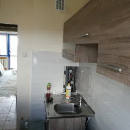 Montaż zlewu w łazience z częściowo położonymi białymi płytkami, widoczne rury instalacji wodnej i szafka pod zlewem z drewnopodobnym dekorem, w tle widoczne pomieszczenie w trakcie remontu.