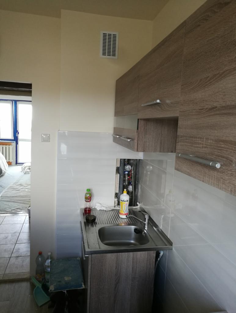 Montaż zlewu w łazience z częściowo położonymi białymi płytkami, widoczne rury instalacji wodnej i szafka pod zlewem z drewnopodobnym dekorem, w tle widoczne pomieszczenie w trakcie remontu.