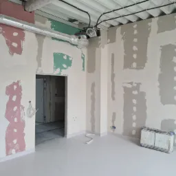 Wnętrze w trakcie prac przygotowawczych do malowania, z widocznymi łatami gipsowymi w kolorach szarym, różowym i zielonym, zabezpieczoną folią podłogą, nową instalacją wentylacyjną pod sufitem...