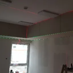 Wnętrze w trakcie montażu płyt kartonowo-gipsowych, widoczne zielone i czerwone linie lasera wyznaczające poziom, okna, drabina, oświetlenie sufitowe.