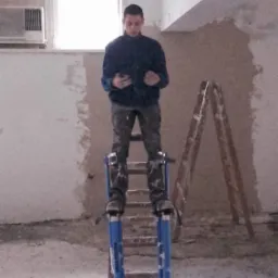 Mężczyzna w niebieskiej kurtce stoi na szczudłach malarskich w surowym wnętrzu, trzymając telefon komórkowy; ściany w trakcie wykańczania, widoczna drabina.