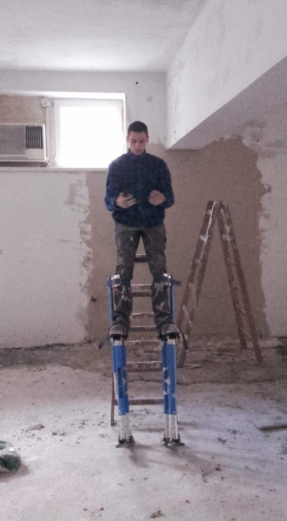 Mężczyzna w niebieskiej kurtce stoi na szczudłach malarskich w surowym wnętrzu, trzymając telefon komórkowy; ściany w trakcie wykańczania, widoczna drabina.