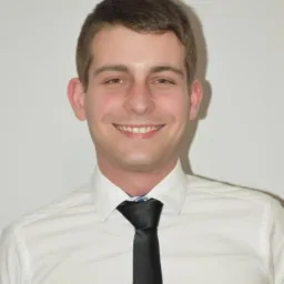 Portret uśmiechniętego mężczyzny w białej koszuli i czarnym krawacie na tle jasnej ściany.