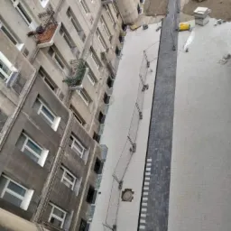 Widok z góry na nowo ułożoną kostkę brukową wzdłuż budynku mieszkalnego, zabezpieczoną tymczasowym ogrodzeniem, z widocznymi elementami infrastruktury podziemnej.