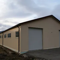 Gotowa hala przemysłowa z panelami warstwowymi, widoczne dwie bramy rolowane, okna wzdłuż bocznej ściany i elementy konstrukcyjne dachu, tło stanowią góry i zachmurzone niebo.