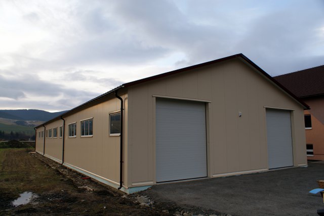 Gotowa hala przemysłowa z panelami warstwowymi, widoczne dwie bramy rolowane, okna wzdłuż bocznej ściany i elementy konstrukcyjne dachu, tło stanowią góry i zachmurzone niebo.