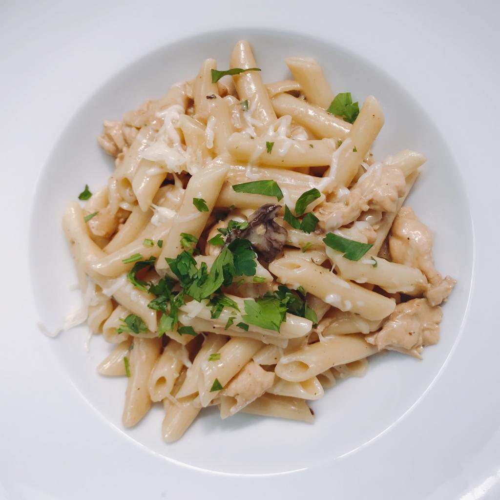 Porcja makaronu penne z kurczakiem w kremowym sosie, posypana świeżą natką pietruszki i startym serem, podana na białym talerzu.
