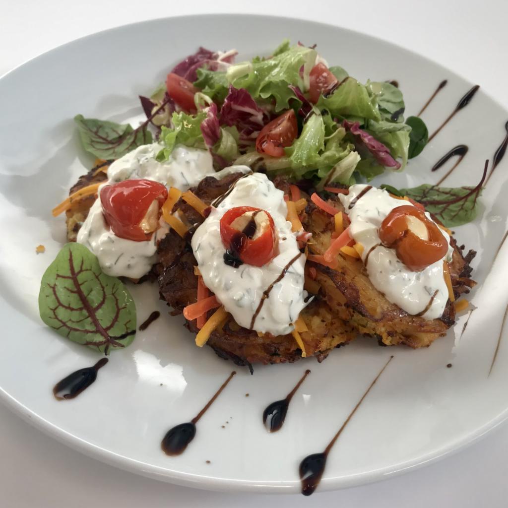 Elegancko podane placki ziemniaczane z sosem ziołowym, pomidorkami cherry i świeżą sałatką na białym talerzu, udekorowane sosem balsamicznym.