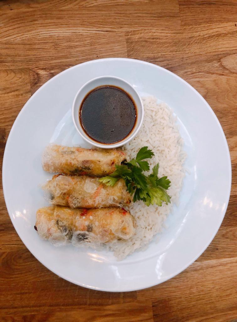 Trzy sajgonki w przezroczystym papierze ryżowym, podane z ryżem, sosem w małej miseczce i gałązką zielonej pietruszki na białym talerzu, ułożone na drewnianym blacie.