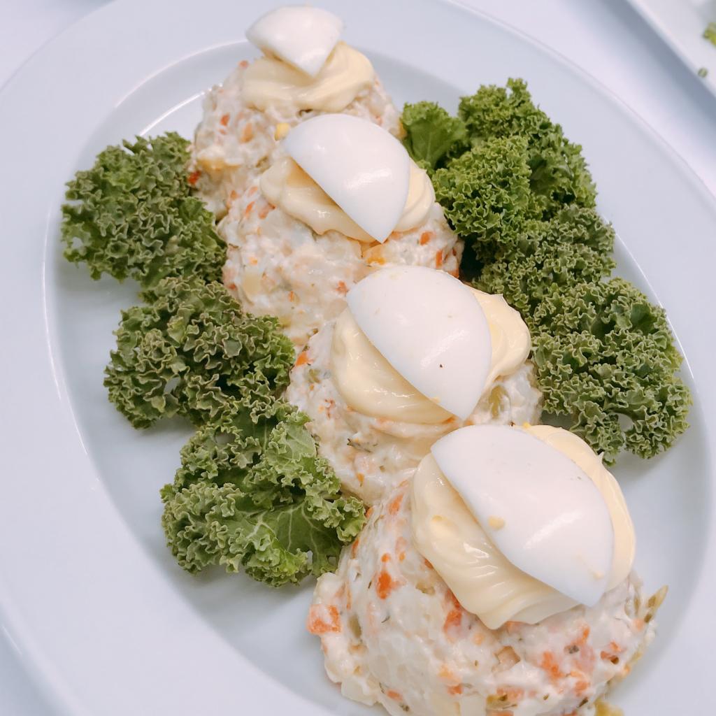 Elegancko podana sałatka jarzynowa z majonezem i jajkiem, udekorowana zielonym kędzierzawym jarmużem, na białym owalnym talerzu.