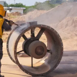 Pracownik w kombinezonie ochronnym czyści strumieniem ściernym duży wentylator przemysłowy na zewnątrz, w tle hałda ziemi i budynek.
