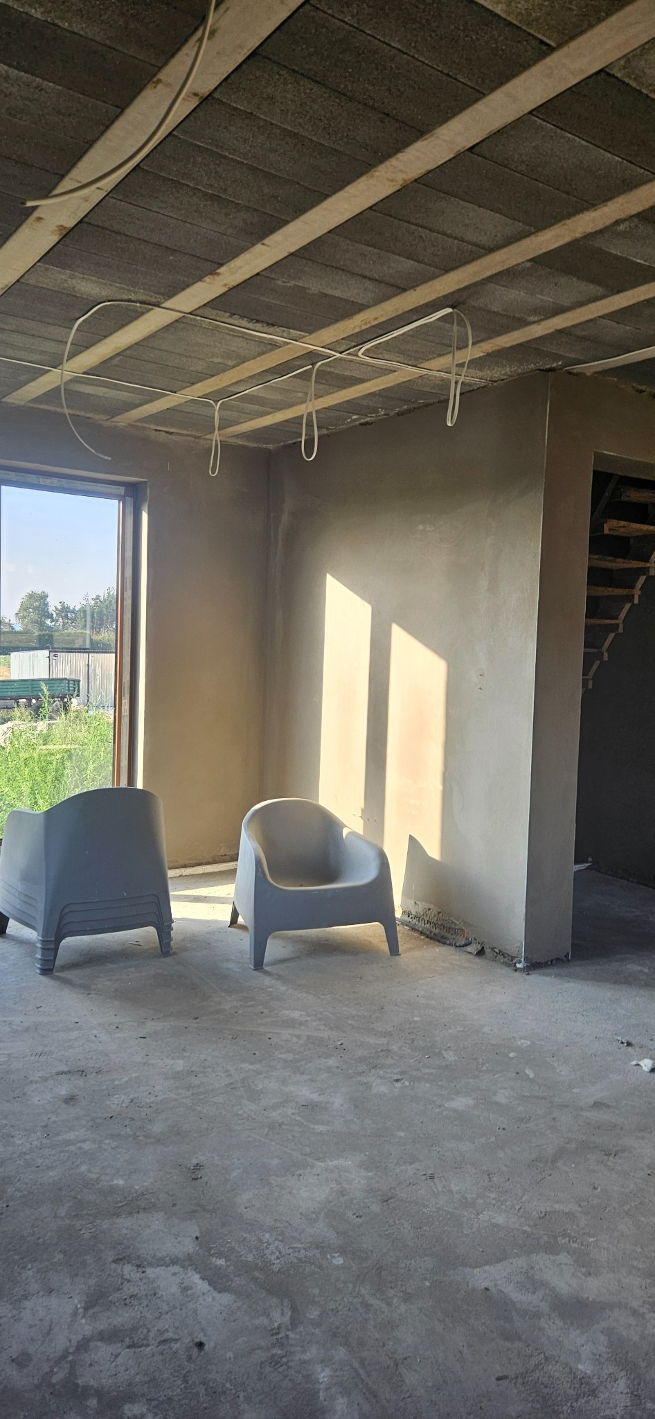 Surowe wnętrze domu w trakcie prac wykończeniowych, z widocznymi świeżo otynkowanymi ścianami, betonowym sufitem z instalacją elektryczną i dwoma plastikowymi krzesłami w pomieszczeniu.