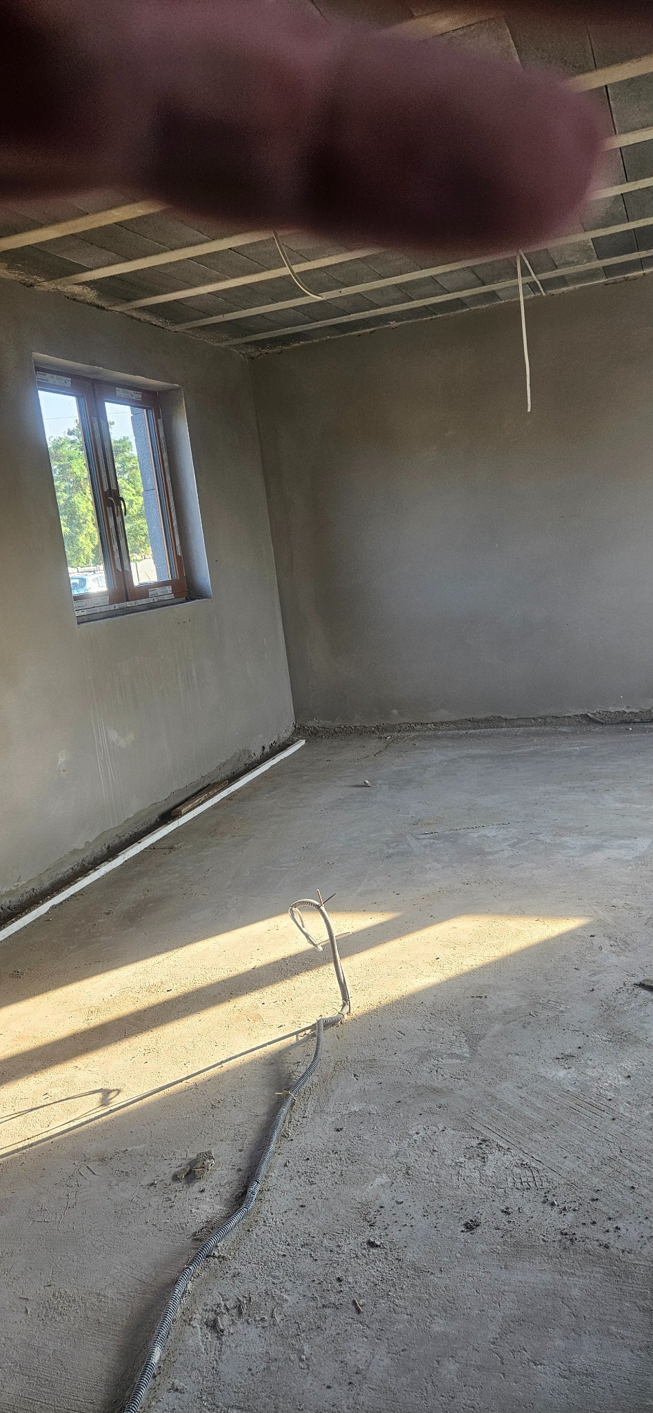 Surowe wnętrze w trakcie prac wykończeniowych: ściany pokryte świeżym tynkiem, widoczne przewody elektryczne na podłodze i drewniane ramy okienne z widokiem na zieleń na zewnątrz.