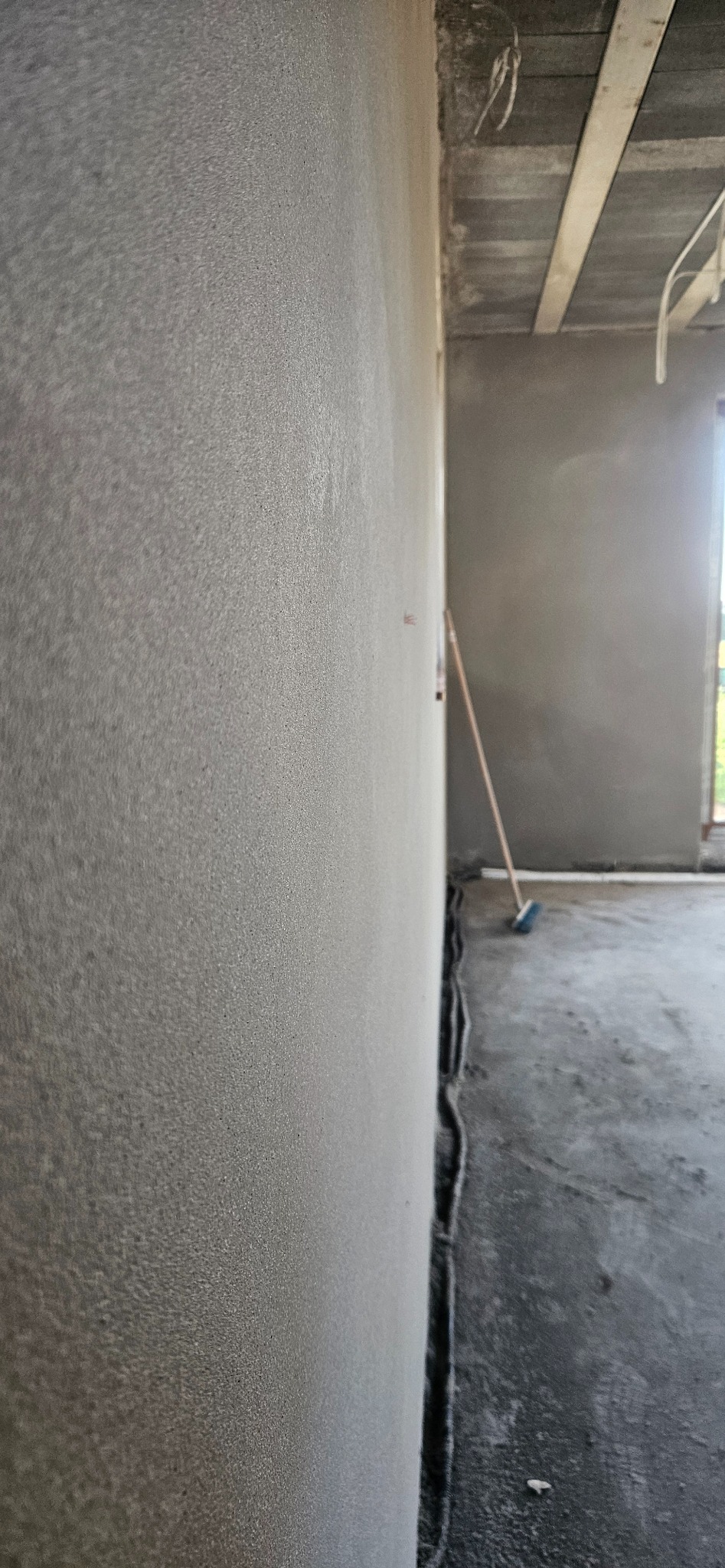 Świeżo położony, jasnoszary tynk maszynowy na ścianie wewnętrznej, widoczne złącze ściany z podłogą zabezpieczone folią, w tle podłoga betonowa i częściowo widoczna błękitna zmiotka.