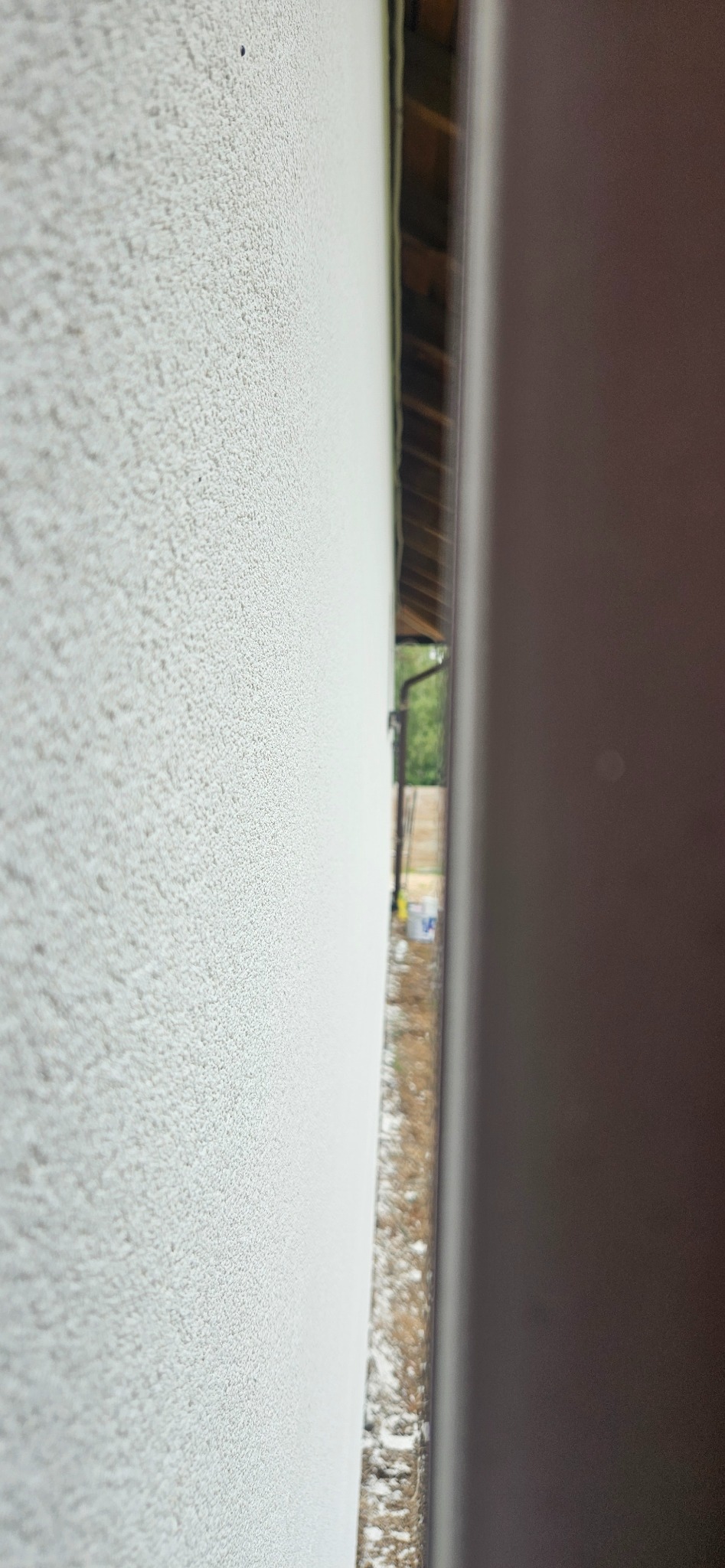 Fragment ściany zewnętrznej z nałożonym tynkiem o wyraźnej fakturze baranka, widoczne elementy konstrukcji dachu i otoczenia przez szczelinę.
