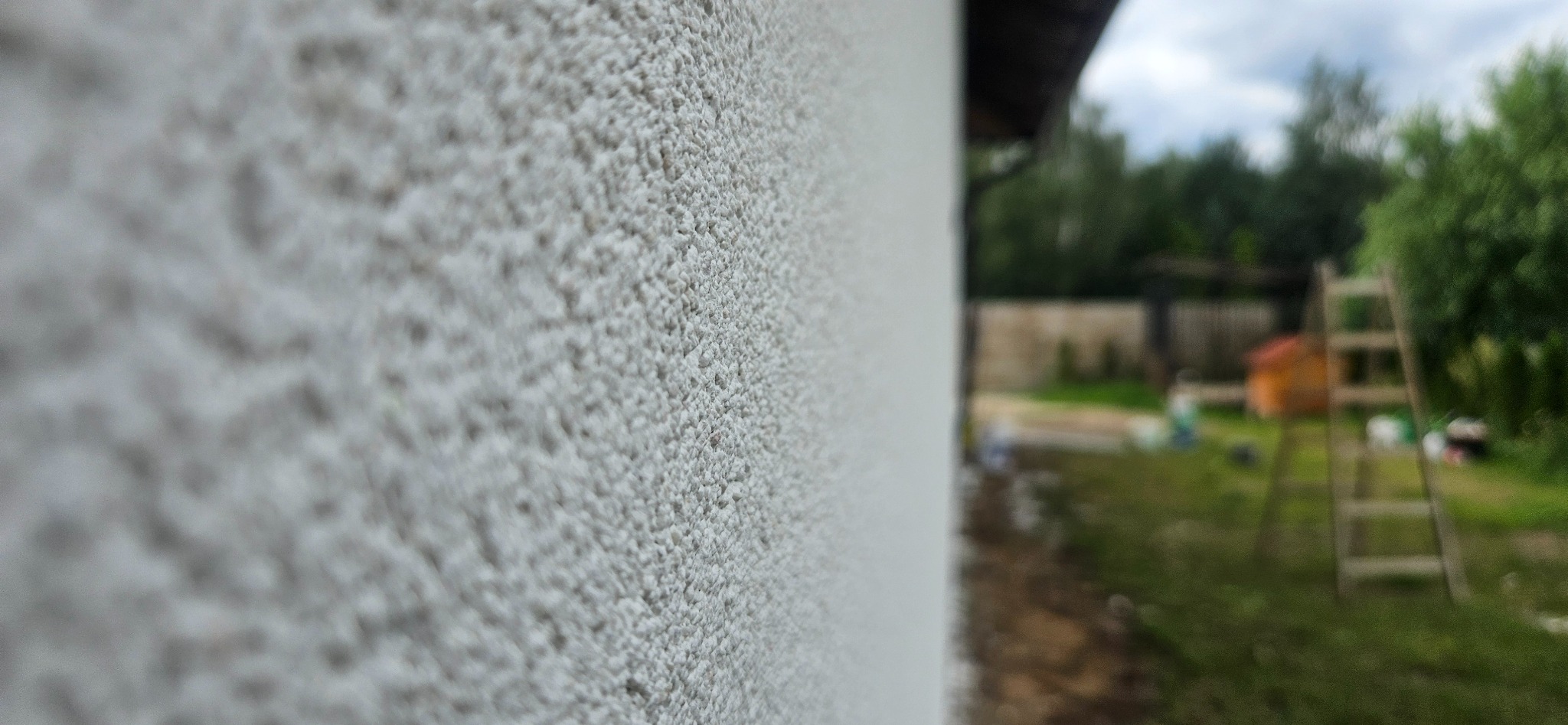 Szczegółowe ujęcie ściany zewnętrznej z tynkiem o fakturze baranka, w tle rozmyty widok ogrodu z drabiną i elementami placu budowy.