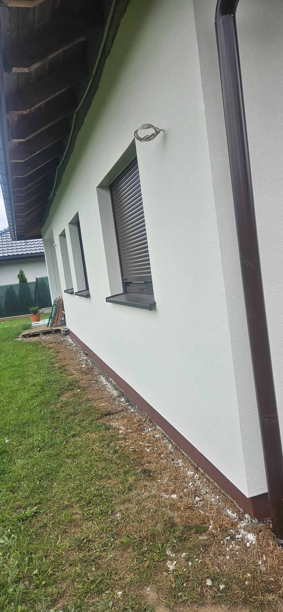 Ściana domu jednorodzinnego po nałożeniu jasnego tynku maszynowego z widocznymi trzema oknami z brązowymi roletami zewnętrznymi, brązową rynną i fragmentem dachu. Wzdłuż ściany widoczne...