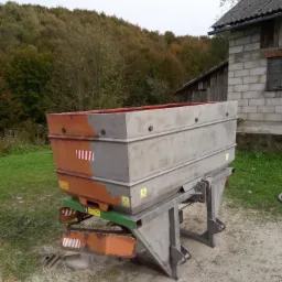 Przyczepa rolnicza z metalową skrzynią, częściowo pomalowana na szaro, z widocznymi znakami ostrzegawczymi, stojąca na piaszczystym podłożu przed budynkiem gospodarczym i zalesionym wzgórzem.