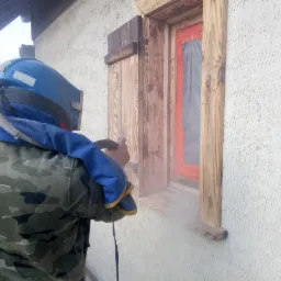 Pracownik w kombinezonie ochronnym czyści drewniane okno z okiennicami za pomocą strumienia ściernego; okno zabezpieczone folią malarską.