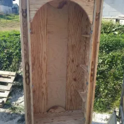 Surowy, drewniany regał z otwartą przestrzenią w kształcie łuku i małymi półkami po boku, widoczny na tle trawy i budynków.