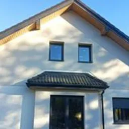 Dom jednorodzinny z jasną, gładką elewacją, ciemnymi oknami i dachem z dachówki ceramicznej w odcieniach szarości, widziany w słoneczny dzień.