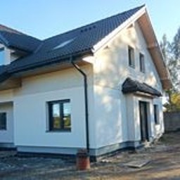 P.W. BANKARO Bożena Bryła - Nowy dom jednorodzinny z jasną, otynkowaną elewacją i czarnymi detalami: okna, dachówka, rynny. Widoczne świeże tynki i przygotowany teren wokół budynku. Częściowo odsłonięte drzewa w tle.