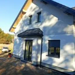 Nowoczesny dom jednorodzinny po ociepleniu elewacji, widoczne okna z roletami zewnętrznymi, jasna elewacja w kontraście z ciemnym dachem, jesienne drzewa w tle, lekki cień na ścianie.