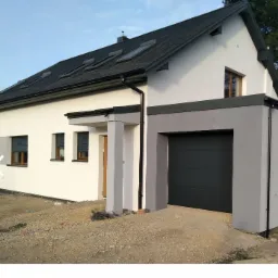 Dom jednorodzinny z garażem po remoncie elewacji, widoczne nowe okna dachowe, szara brama garażowa i świeżo pomalowana biała elewacja, teren wokół domu częściowo utwardzony.