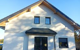 Dom jednorodzinny z jasną, gładką elewacją, ciemnymi oknami i dachem z dachówki ceramicznej w odcieniach szarości, widziany w słoneczny dzień.