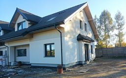 Nowy dom jednorodzinny z jasną, otynkowaną elewacją i czarnymi detalami: okna, dachówka, rynny. Widoczne świeże tynki i przygotowany teren wokół budynku. Częściowo odsłonięte drzewa w tle.