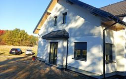 Nowoczesny dom jednorodzinny po ociepleniu elewacji, widoczne okna z roletami zewnętrznymi, jasna elewacja w kontraście z ciemnym dachem, jesienne drzewa w tle, lekki cień na ścianie.