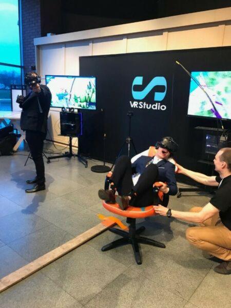 Mężczyzna w garniturze siedzi w krześle obrotowym z założonym zestawem VR, podczas gdy druga osoba pomaga mu utrzymać równowagę, w tle widać logo VR Studio i monitory.