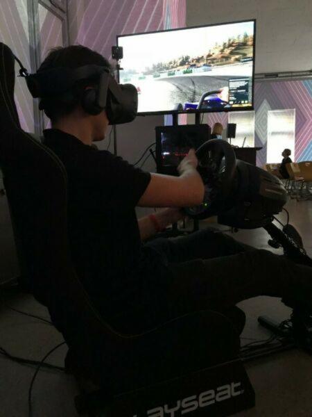 Osoba w czarnym ubraniu, siedząca w fotelu Playseat, używa zestawu VR i kierownicy do symulacji wyścigów samochodowych na dużym ekranie.