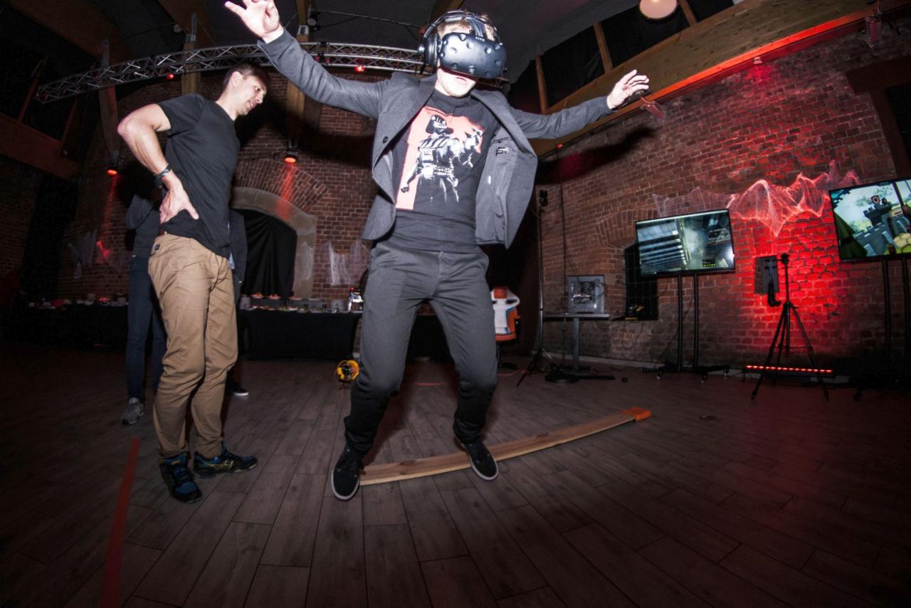 Mężczyzna w garniturze i goglach VR skacze na desce podczas eventu, w tle ceglana ściana i monitory z grami.