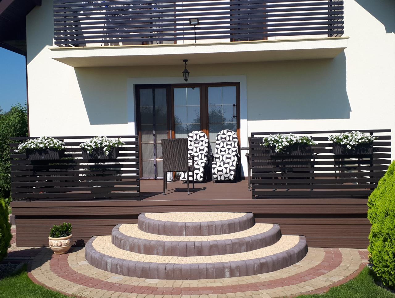 Elegancki taras z drewnianą podłogą, balustradą z poziomych listew, donicami z białymi kwiatami, schodami z kostki brukowej i dwoma krzesłami z wzorzystymi poduszkami, widok na białą elewację domu...