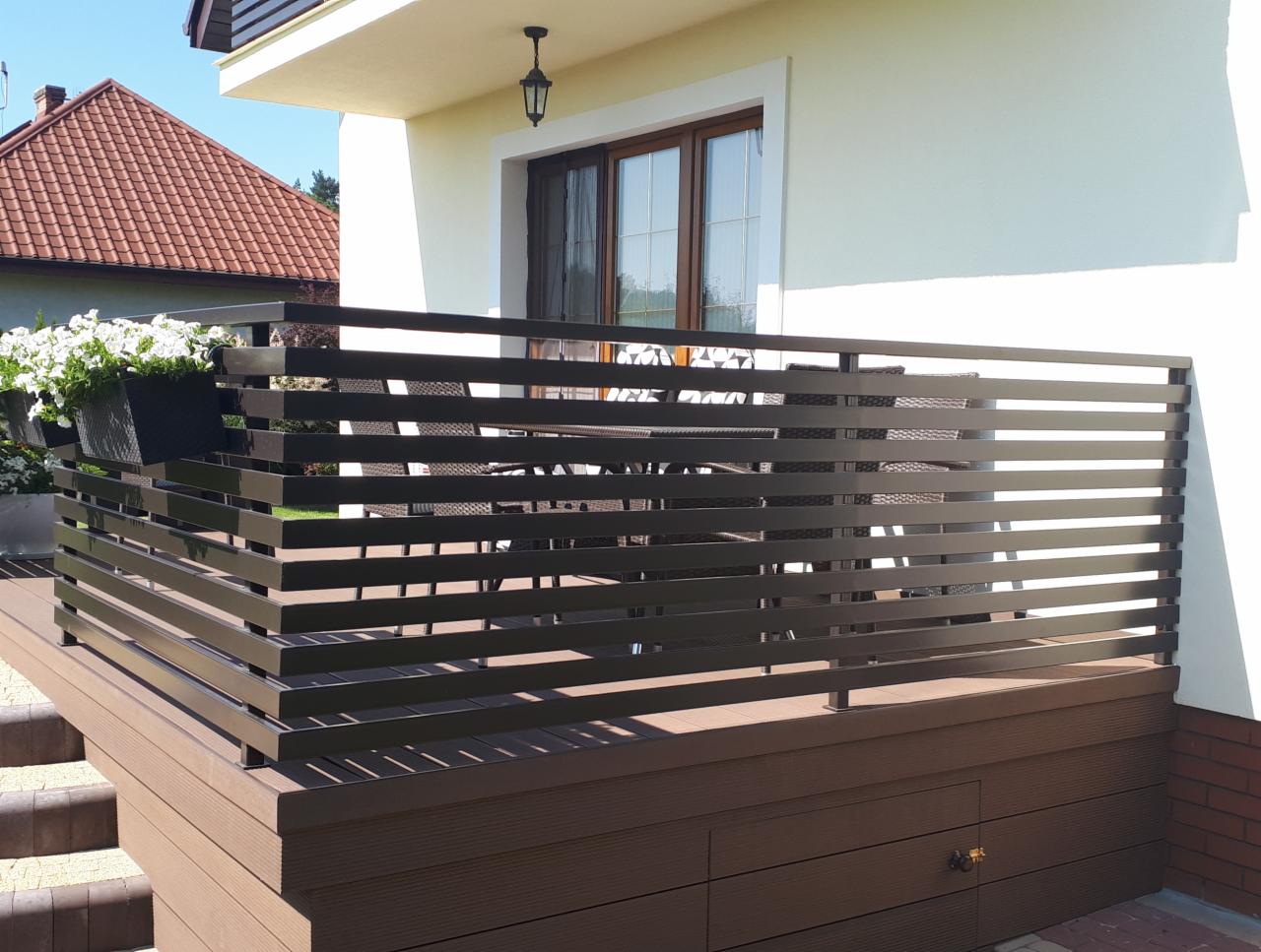 Nowoczesna balustrada balkonowa z poziomych, brązowych profili metalowych, widok z dołu na taras z meblami ogrodowymi i kwiatami w donicach.