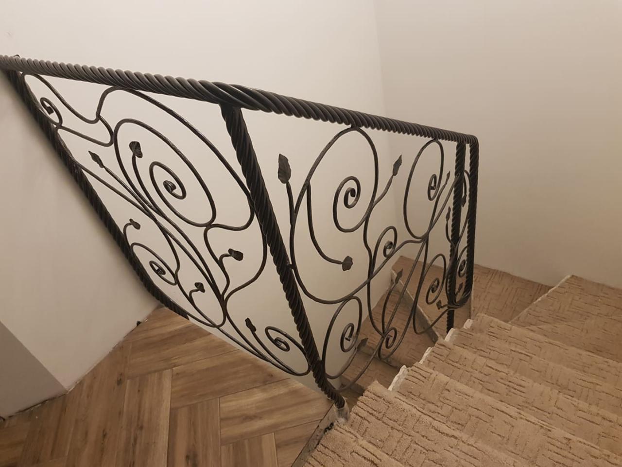 Kute balustrady schodowe z motywem roślinnym, czarne, na tle białej ściany i drewnopodobnych płytek podłogowych oraz schodów pokrytych wykładziną.