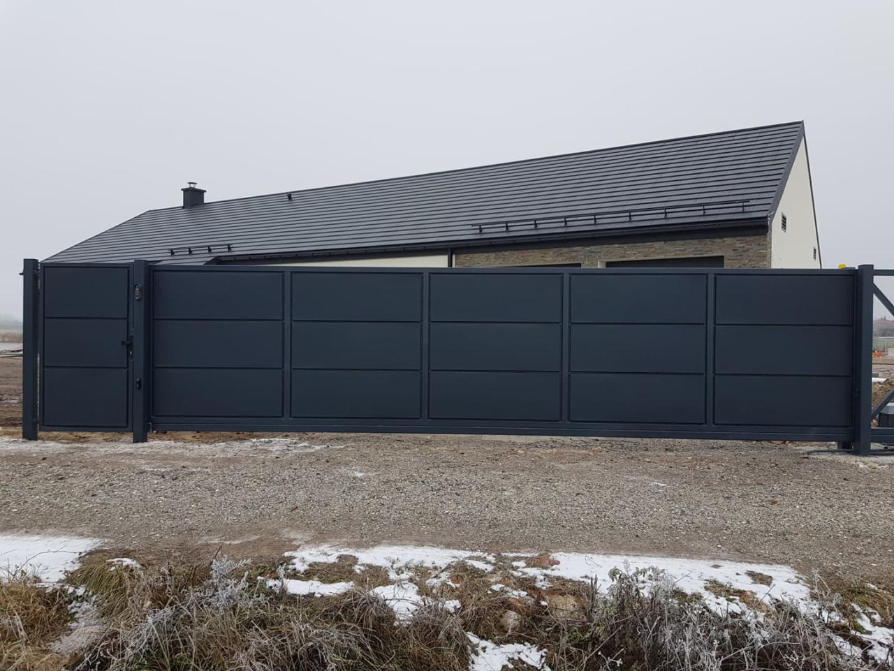 Nowoczesna, przesuwna brama wjazdowa w kolorze grafitowym, z furtką po lewej stronie, na tle domu z ciemną dachówką i pochmurnego nieba.