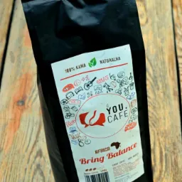 Czarna paczka kawy ziarnistej You.Cafe Africa Bring Balance, leżąca na drewnianym blacie z widocznym logo i informacjami o produkcie.