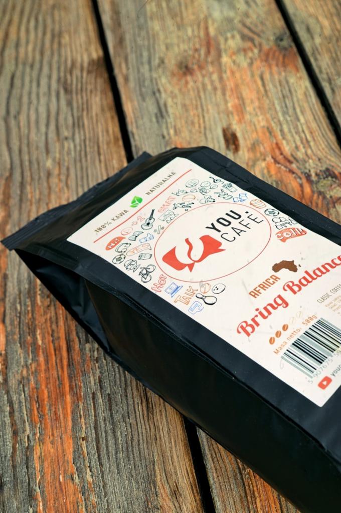 Czarna paczka kawy ziarnistej 'You Cafe Bring Balance Africa' leżąca na drewnianym blacie z widocznym logo i informacjami o produkcie.